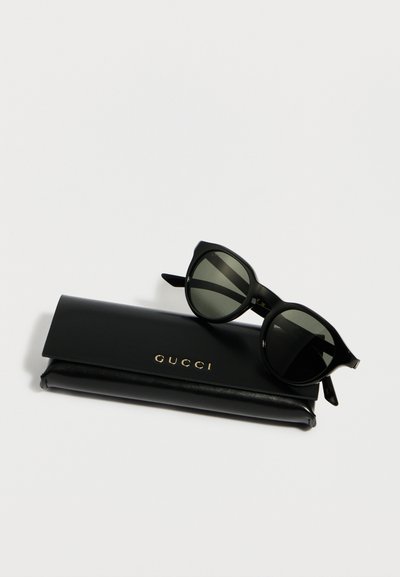 Μαύρα γυαλιά ηλίου σε σχήμα cat-eye που ακουμπούν σε μαύρη δερμάτινη θήκη με λογότυπο Gucci, πάνω σε λευκό φόντο.