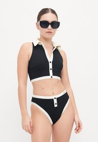Black ribbed bikini set with white collar and button accents; cropped top and high-waisted bottom with contrasting trim. Black sunglasses.