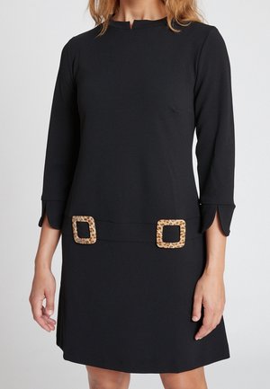 Kobieta ubrana w czarną sukienkę z rękawami 3/4, małym rozcięciem na przodzie dekoltu oraz dwoma kwadratowymi klamrami w panterkę na talii.