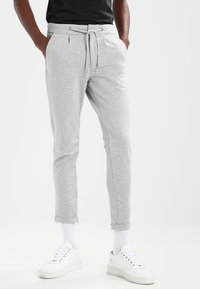 Pantalones de chándal gris claro hechos de tela suave, con cintura ajustable mediante cordón, puños ceñidos y bolsillos laterales. Combinados con zapatillas blancas.