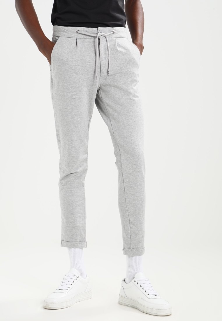Pantalones de chándal gris claro hechos de tela suave, con cintura ajustable mediante cordón, puños ceñidos y bolsillos laterales. Combinados con zapatillas blancas.