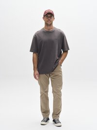 Grijze oversized T-shirt, beige broek met rechte pijpen en zwart-witte gestreepte sneakers. Rode pet met witte tekstdetails. Effen achtergrond.