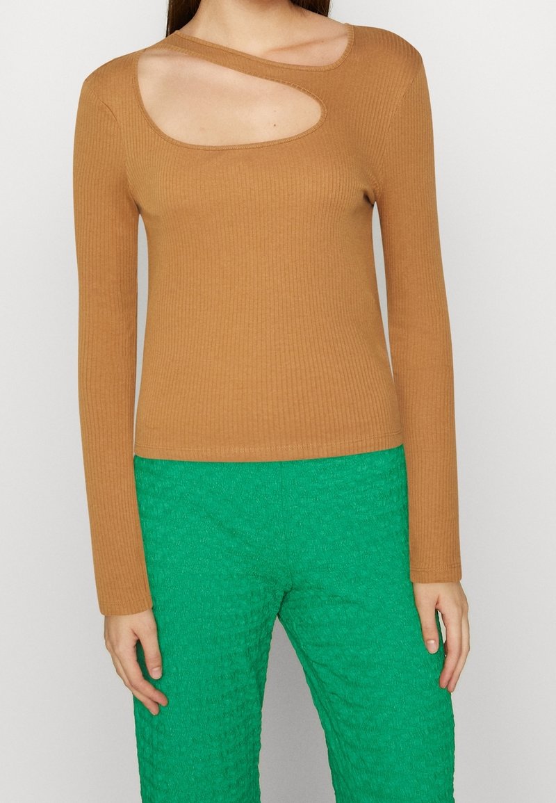 Ruskea ribattu pitkähihainen paita, jossa on ainutlaatuinen leikattu muoto kaula-aukossa, yhdistettynä teksturoituihin vihreisiin housuihin.