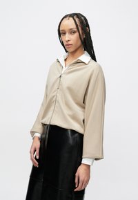 Μπεζ τοπ με φερμουάρ, λευκό γιακά, μακριά μανίκια και άνετη γραμμή. Συνδυασμένο με μαύρη γυαλιστερή δερμάτινη φούστα.