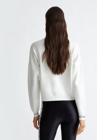 Weißer, kurzer Pullover mit langen Ballonärmeln, strukturiertem Stoff und gerippten Bündchen. Kombiniert mit schwarzen Leggings mit Spitzenakzenten.