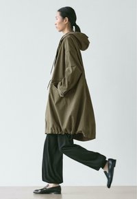 Olivgrüne Kapuzenjacke mit weiter Passform, Tunnelzug in der Taille und großen Seitentaschen, getragen über weiten schwarzen Hosen und flachen Schuhen.