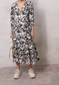 Kerman midimekko, jossa on musta ja beige kuviointi, 3/4-hihat ja kerrostettu helma. Yhdistettyt paksut beige-kengät ja valkoinen laukku.