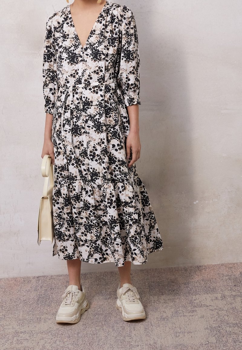 Kerman midimekko, jossa on musta ja beige kuviointi, 3/4-hihat ja kerrostettu helma. Yhdistettyt paksut beige-kengät ja valkoinen laukku.