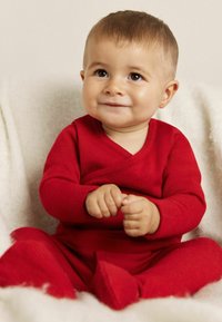 Punainen neuleasu, jossa on kietaisumalli, pitkät hihat ja sopivat jalkapohjat. Lapsi istuu kevyellä, teksturoidulla peitteellä.