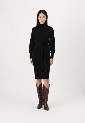 Schwarzes Strickkleid mit hohem Kragen, langen Ärmeln und einem Taillengürtel. Tragen mit braunen gemusterten Cowboy-Stiefeln. Glatte Textur.