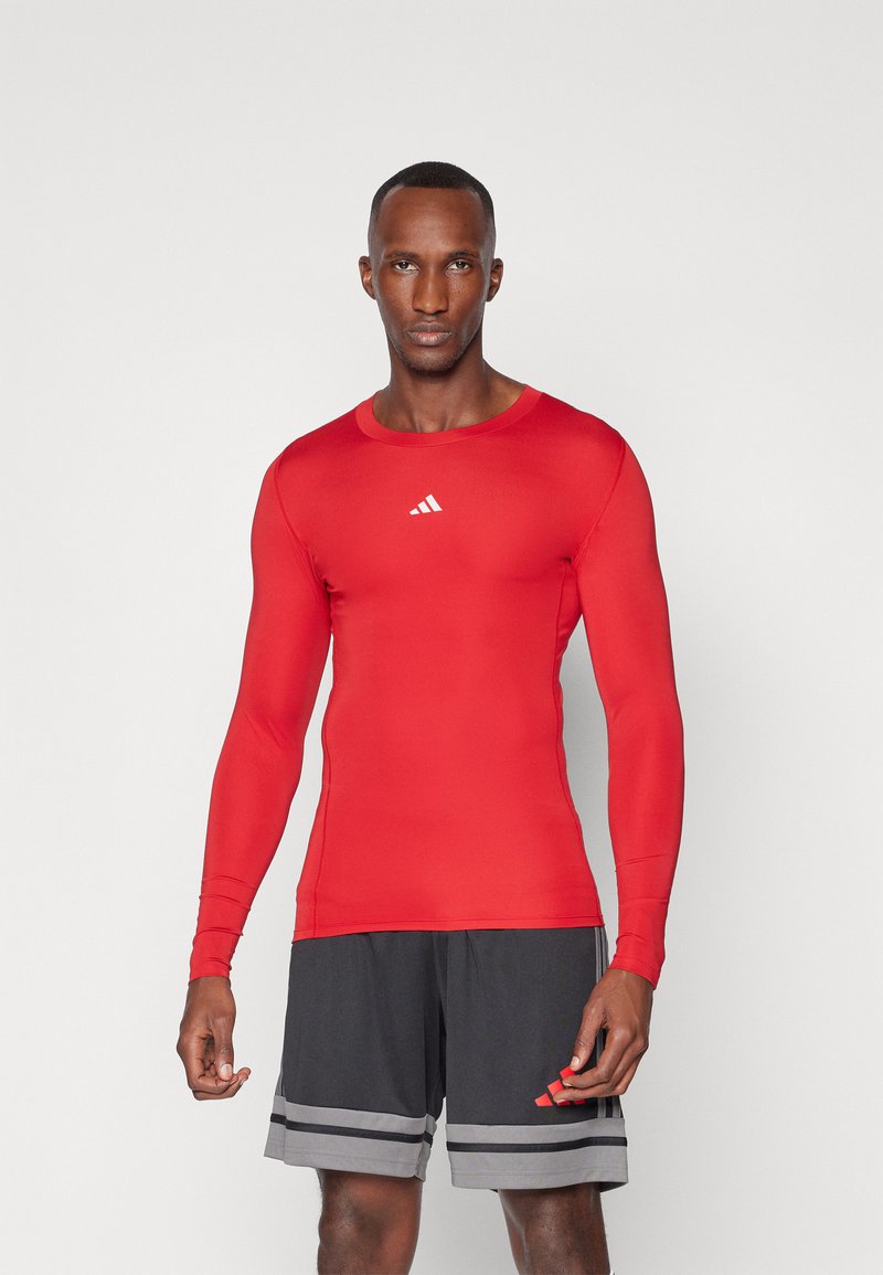 Rød langærmet træningsshirt med rund hals, lavet af strækbart stof; har et hvidt Adidas-logo på brystet og en tætsiddende design.