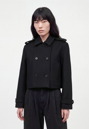 Un mannequin aux longs cheveux foncés porte un manteau court noir croisé et un pantalon noir ample, et se tient devant un fond clair uni.