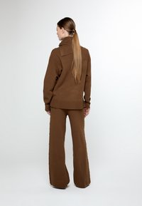 Kvinna med långt hår i en brun höghalsad tröja och vida byxor med sidoknappar, stående vänd bort från kameran mot en enfärgad bakgrund.