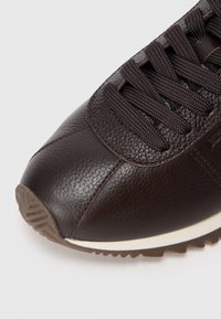 Zapatilla de cuero marrón con acabado texturizado, con cordones oscuros, una entresuela blanca contrastante y una suela de goma para mayor tracción.