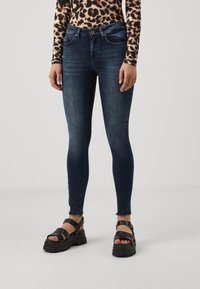Femme portant un jean skinny bleu foncé, un haut à manches longues imprimé léopard et des sandales à plateforme noires, debout sur un fond clair uni.