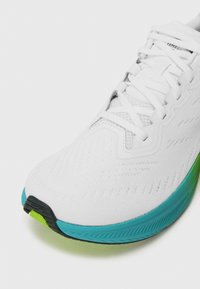 Baskets blanches avec un dessus en tricot texturé, présentant des accents turquoise vif et vert sur la semelle et un capuchon de talon noir.