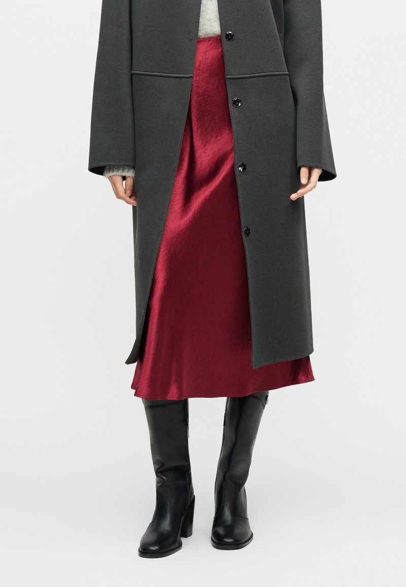 Άτομο που φοράει γκρι μακρύ παλτό πάνω από κόκκινη σατέν φούστα με μαύρες ψηλές μπότες μέχρι το γόνατο, σε απλό φόντο.