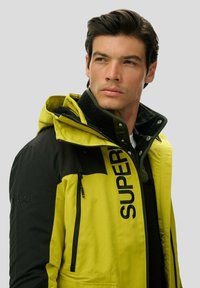 Lysegul og sort jakke med lynlås og høj krave, med "SUPER" logo foran og detaljer med tekstureret stof.