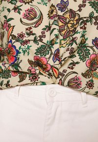 Cremefarbene Bluse mit floralem Muster, die lebendige Pink-, Blau- und Grüntöne zeigt, an der Taille verknotet über weißen Denim-Hosen.
