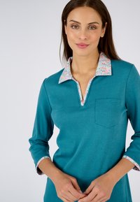 Teal langemouw polo shirt met een bloemenpatroon op de kraag en manchetten; inclusief een klein borstzakje. Glad textuur en een relaxte pasvorm.