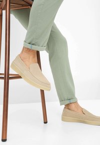 Scarpe beige slip-on con una finitura in camoscio liscio, dotate di una suola in gomma di colore chiaro. Indossate con pantaloni verde chiaro arrotolati.