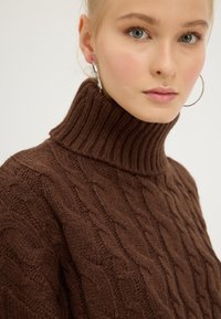 Brun kabelstrikket genser med høy ribbestrikket hals. Stoffet ser teksturert og tykt ut, og viser et tydelig mønster gjennom hele genseren.