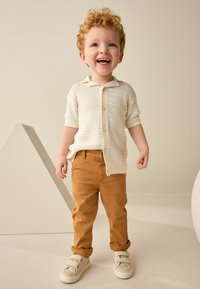 Cardigan en tricot beige clair avec des boutons, associé à un pantalon beige et des baskets grises. Enfant aux cheveux bouclés sourit sur fond neutre.