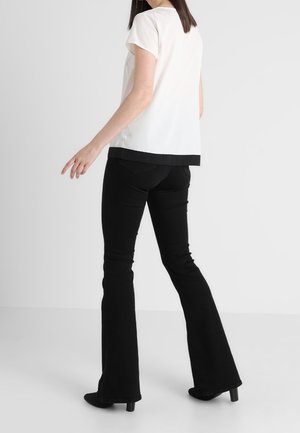 Kobieta w czarnych rozszerzanych spodniach i białym krótkim topie z czarną lamówką, stojąca na jednolitym białym tle.