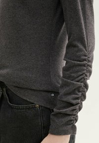 Maglietta grigia a maniche lunghe con dettagli arricciati sulle maniche, realizzata in un tessuto morbido e testurizzato. Etichetta logo laterale ben visibile e vestibilità comoda.