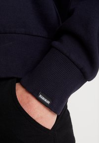 Sudadera de color marino exhibida, con puños acanalados y una etiqueta de marca. La tela parece suave; lleva puesta sobre ropa negra con una textura lisa.