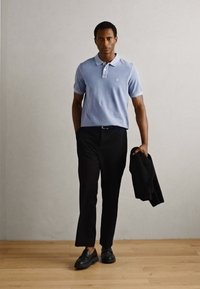 Camiseta polo azul claro con cuello, pantalones negros y chaqueta negra. La camiseta presenta un pequeño logo; zapatos negros completan el atuendo.