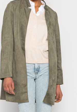 Giacca in suede verde oliva con una vestibilità rilassata, apertura frontale e maniche lunghe, indossata sopra un maglione beige chiaro e jeans azzurri chiari.