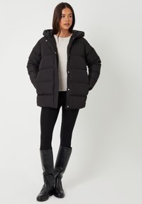 Sort pufferjakke med hætte, der har et firkantet design, syede kvadrater og lynlås. Båret over en lys top og sorte leggings.