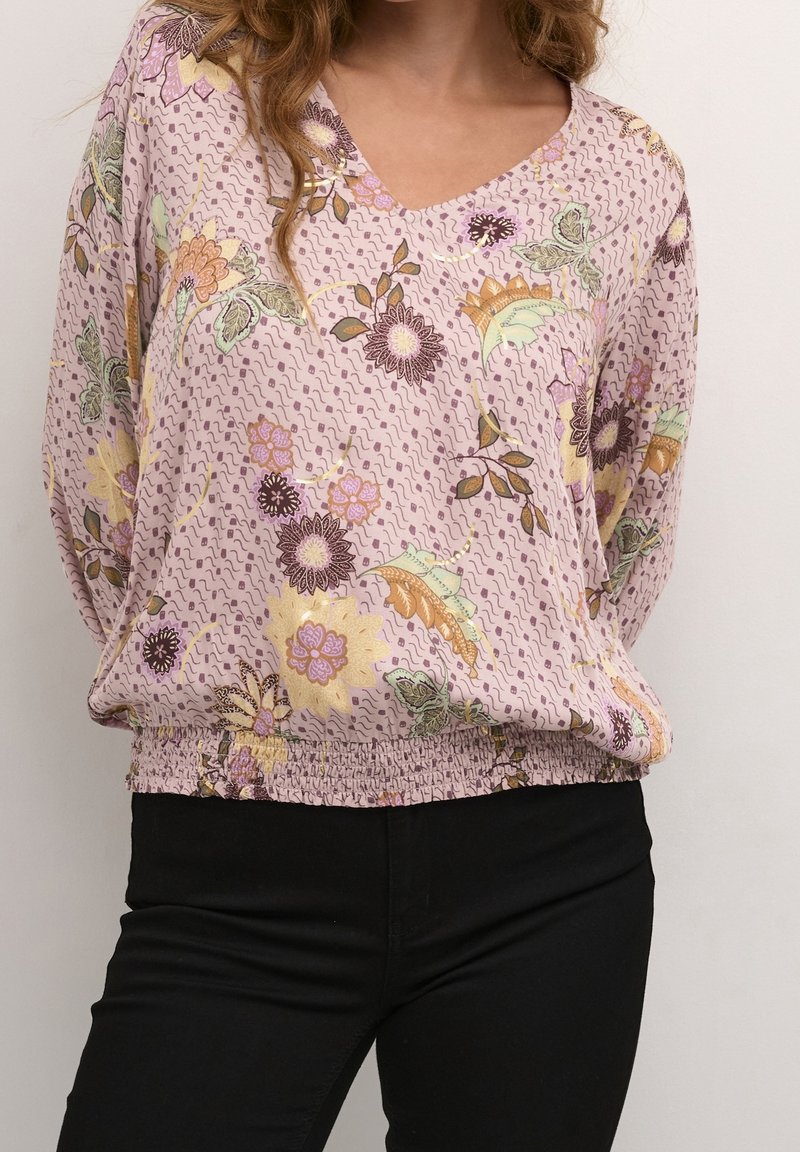 Femme portant un chemisier floral violet clair avec une taille smockée et un pantalon noir, debout devant un fond blanc uni.