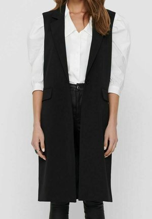 Czarna bezrękawnik o dopasowanym kroju, z pojedynczym zapięciem na guzik i dwiema kieszeniami z przodu, noszony na białej koszuli.