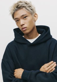 Tengerészkék, oversized kapucnis pulóver nagy kapucnival, bordás mandzsettákkal és varrás nélküli dizájnnal, fehér ing fölött viselve, kezek keresztbe tett állapotban. Simává textúrával.