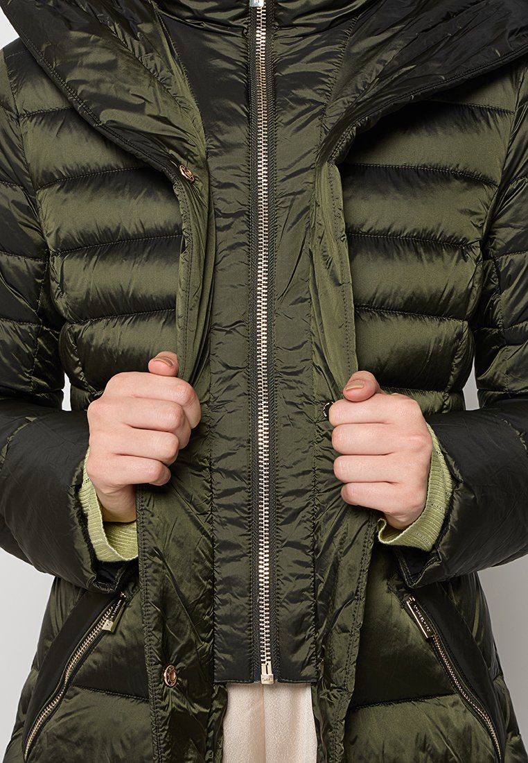 Grüne gesteppte Daunenjacke mit doppeltem Reißverschluss, Hände ziehen die Front auf, geschichtet über hellgrüne Ärmel und darunter ein beiges Kleidungsstück.