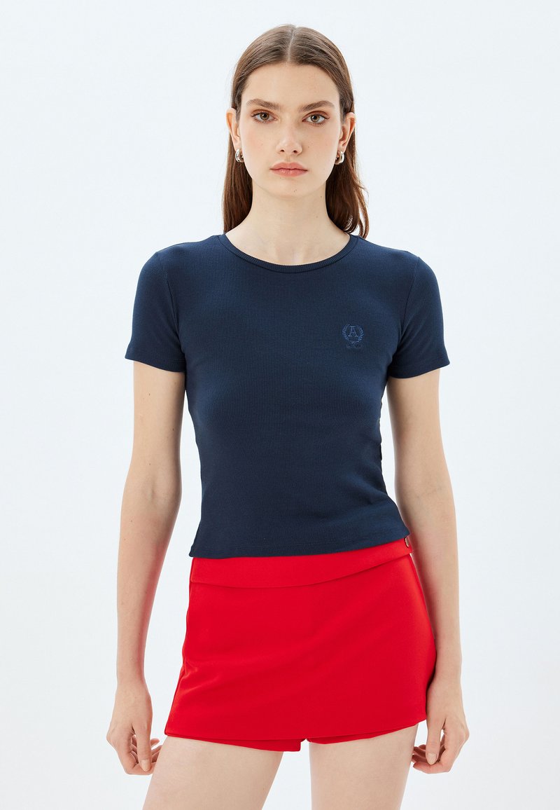 Marineblå ribbet t-shirt med korte ærmer, rund hals og lille broderet logo på venstre bryst, parret med en rød nederdel.