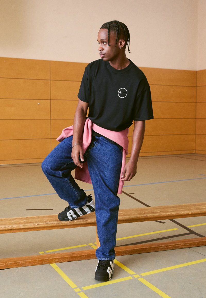 Schwarzes T-Shirt mit weißem Logo, rosa Sweatshirt um die Taille gebunden, blaue Jeans, schwarze Schuhe mit weißen Akzenten, stehend auf einer Holzbank in einem Fitnessstudio.