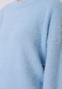 Svetlo modra majica z dolgimi rokavi iz mehke, teksturirane tkanine. Ima okrogel izrez in sproščen kroj z puhastim, udobnim videzom.