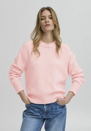 Pull en maille rose avec un col rond et des poignets côtelés, arborant un petit logo blanc. Assorti avec un jean en denim bleu. Texture douce.