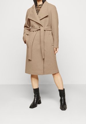 Femme portant un manteau beige ceinturé mi-long, des bottines noires et un haut à motifs, debout devant un fond clair.