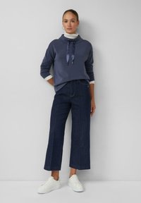 Marineblauer Pullover mit Schlauchkragen und Satinkordeln, kombiniert mit weit geschnittenen, dunklen Denim-Hosen und weißen Turnschuhen.