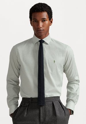 Man wearing light checkered dress shirt with dark knit tie and gray pleated trousers, standing with hands in pockets against white background.