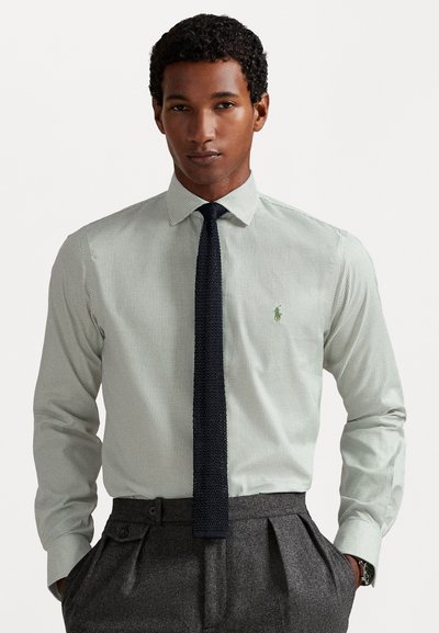 Uomo che indossa una camicia a quadri leggeri con una cravatta di maglia scura e pantaloni grigi plissettati, in piedi con le mani in tasca su uno sfondo bianco.