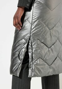Metallisch graue Daunenjacke mit einem Chevron-Steppmuster. Mit seitlichem Schlitz und Druckknöpfen ausgestattet. Über schwarzem Streifenstoff getragen.