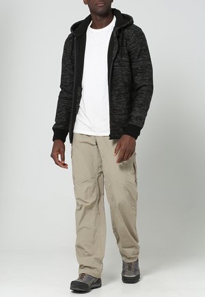 Felpa nera e grigia con cordini, t-shirt bianca sotto, pantaloni cargo beige chiaro e scarponi da trekking neri.