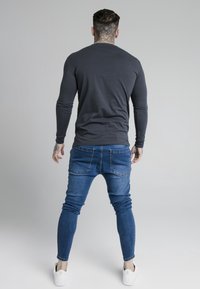 Long-sleeve gray shirt, fitted design, paired with blue slim-fit jeans and white sneakers. Smooth fabric, casual look, and accented with tattoos.
