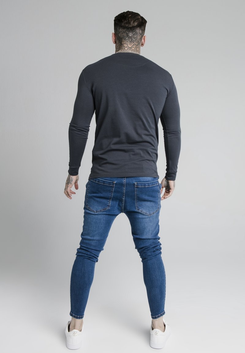 Long-sleeve gray shirt, fitted design, paired with blue slim-fit jeans and white sneakers. Smooth fabric, casual look, and accented with tattoos.