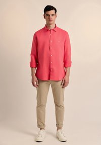 Camisa de botones coral ligera con mangas enrolladas, combinada con pantalones beiges y zapatillas blancas, con un pequeño logo bordado.